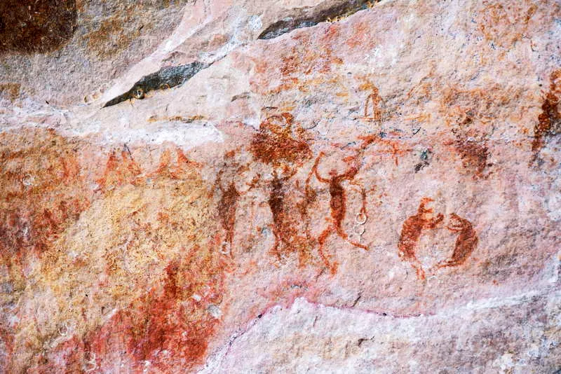 Dos personajes de perfil, en posicin de sentadillas, discuten; uno de ellos muestra sus fauces abiertas con colmillos. Otros cazadores guerreros bailan a su lado. 